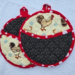 Puede incluir: Dos agarraderas redondas acolchadas con ribete y lazo para colgar rojos. La sección superior presenta un estampado de gallo sobre un fondo crema, mientras que la sección inferior es negra con un estampado floral blanco. Lunares blancos acentúan el ribete rojo.
