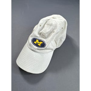 May include: A white baseball cap featuring a centered oval patch with a dark blue background and a yellow letter M embroidered in the center. The cap has a curved brim, stitched eyelets on the crown, and a structured front panel design.
