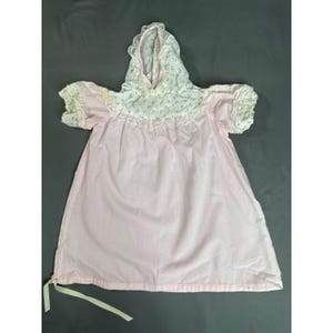 Pode incluir: Um vestido com capuz em rosa claro, apresentando um jugo branco com um pequeno padrão floral rosa. A peça inclui mangas curtas com acabamento em renda franzida, um capuz a condizer com renda e uma fita clara na bainha inferior.