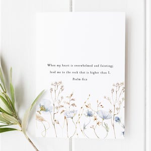 Puede incluir: Impresión rectangular blanca con un diseño floral en acuarela. Flores azules y beige adornan la parte inferior. El texto dice: «Cuando mi corazón desfallece, llévame a la roca que es más alta que yo. Salmo 61:2.»