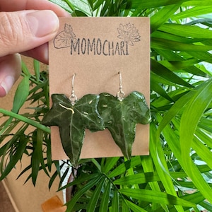 Puede incluir: Un par de pendientes de hoja verde con ganchos plateados. Los pendientes están en una tarjeta marrón con el texto "MOMOCHARI" impreso en ella.