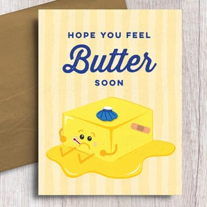 May include: A greeting card with a cartoon butter character and the words "Hope You Feel Butter Soon". The card has a yellow and white striped background and is accompanied by a brown envelope.