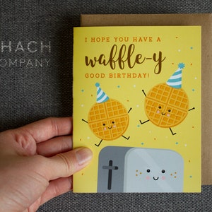 May include: A yellow birthday card with a waffle-themed design. The card features two smiling waffles wearing party hats and a toaster with a happy face. The text on the card reads "I hope you have a waffle-y good birthday!"
