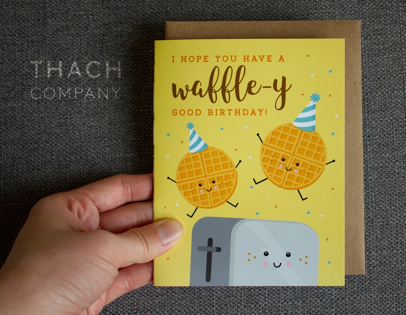 May include: A yellow birthday card with a waffle-themed design. The card features two smiling waffles wearing party hats and a toaster with a happy face. The text on the card reads "I hope you have a waffle-y good birthday!"