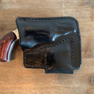 Pode incluir: Um coldre de couro preto com uma pequena pistola dentro. A pistola tem um cabo de madeira de cor marrom avermelhada.