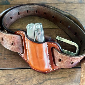 May include: A brown leather belt with a holster for a multi-tool. The holster is attached to the belt and has a snap closure. The multi-tool is a silver Leatherman.