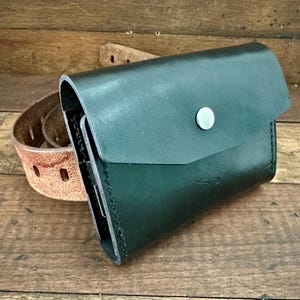 May include: A dark green leather pouch with a flap secured by a silver snap. The pouch is next to a brown leather belt with a buckle. The items are on a wooden surface.