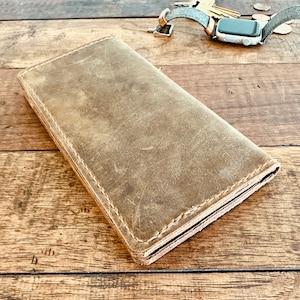 Puede incluir: Una cartera de cuero marrón desgastado con costuras visibles en los bordes. La cartera rectangular parece estar cerrada. Llaves, monedas y un reloj inteligente con una correa gris son visibles en el fondo sobre una superficie de madera.