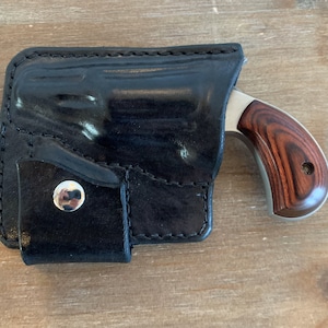 Pode incluir: Um coldre de couro preto com um bot&atilde;o prateado e uma pequena pistola prateada com cabo de madeira.