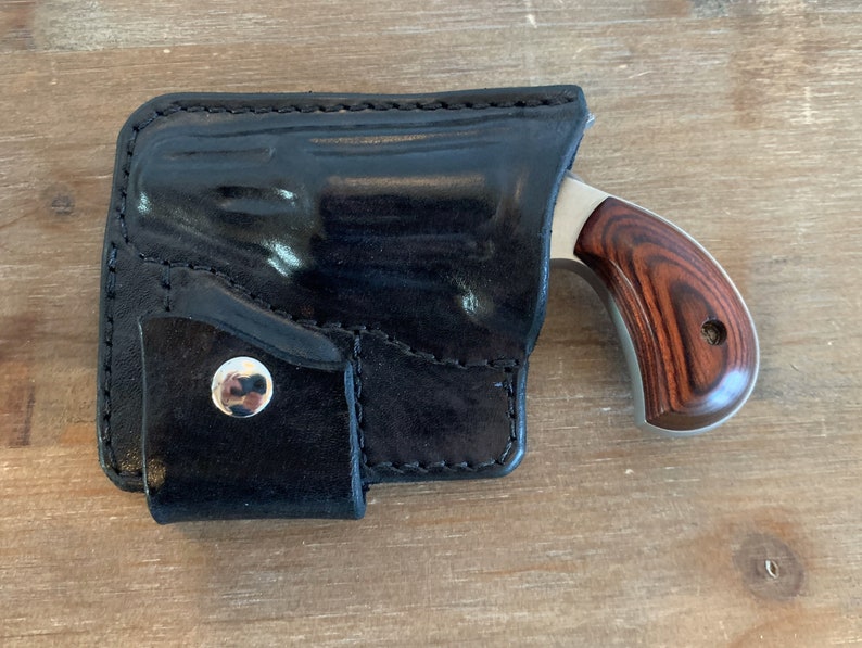 Pode incluir: Um coldre de couro preto com um bot&atilde;o prateado e uma pequena pistola prateada com cabo de madeira.