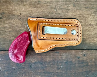 Könnte beinhalten: Ein braunes Lederholster mit einem Metallclip und einem roten Kunststoffpistolengriff.