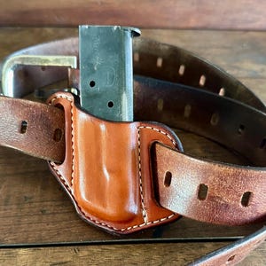 Może przedstawiać: Brązowy skórzany pasek z przymocowaną brązową skórzaną kaburą. Kabura zawiera magazynek.