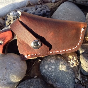 May include: A brown leather holster with white stitching and a silver button. A small wooden-handled firearm is partially visible inside the holster.