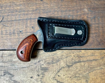 Könnte beinhalten: Eine kleine, silberne Pistole mit einem Holzschaft befindet sich in einem schwarzen Lederholster. Das Holster hat einen Metallclip.