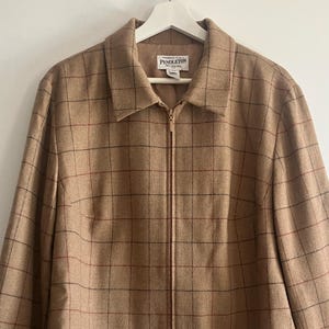 May include: A tan jacket with a dark brown and rust-colored grid pattern. The jacket features a full-length front zipper, a pointed collar, and long sleeves. A white label inside the collar displays the text Pendleton in black lettering.