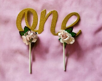 Könnte beinhalten: Goldener Glitzer-Kuchentopper "eins" mit rosa und weißen Blumen auf weißen Stielen.