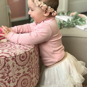 May include: A young child wearing a pink long-sleeved shirt and a white tulle skirt with a gold waistband. The child is wearing a flower crown with pink and white flowers. The child is standing next to a pink and white patterned ottoman.