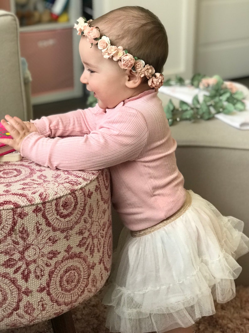May include: A young child wearing a pink long-sleeved shirt and a white tulle skirt with a gold waistband. The child is wearing a flower crown with pink and white flowers. The child is standing next to a pink and white patterned ottoman.