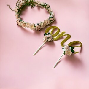 May include: A gold glitter "one" cake topper with white flowers and green leaves, and a white flower crown with green leaves.