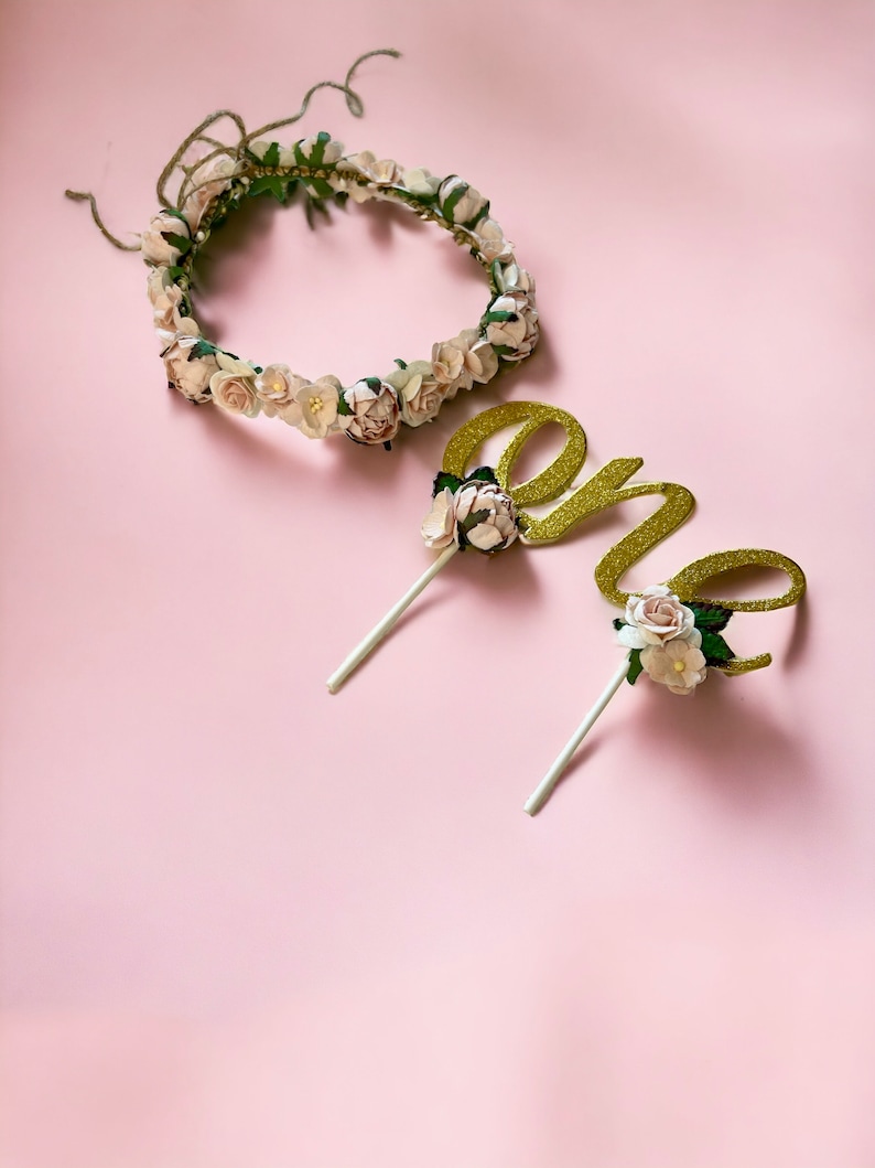 May include: A gold glitter "one" cake topper with white flowers and green leaves, and a white flower crown with green leaves.