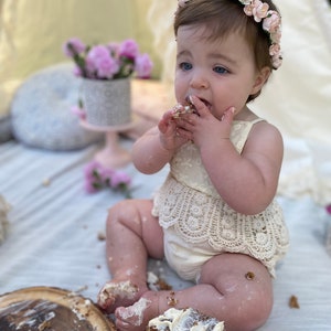 May include: A baby girl wearing a white dress with lace trim and a pink flower crown is sitting on a white blanket and eating cake. The cake is on a wooden plate and there are crumbs on the blanket.