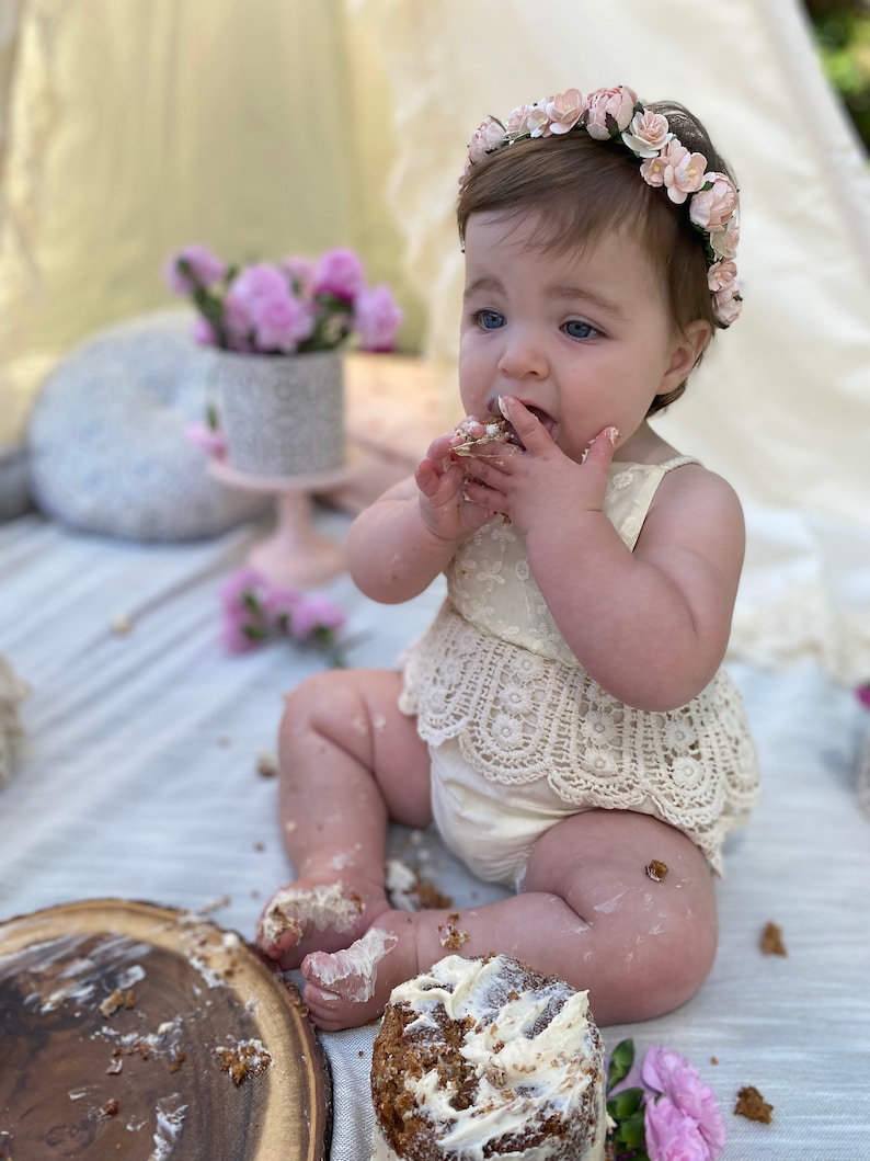 May include: A baby girl wearing a white dress with lace trim and a pink flower crown is sitting on a white blanket and eating cake. The cake is on a wooden plate and there are crumbs on the blanket.