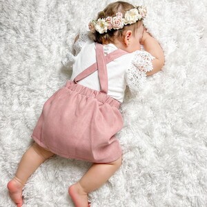 May include: A baby girl wearing a pink skirt and a white shirt with straps. She is wearing a flower crown and is lying on a white fluffy rug.