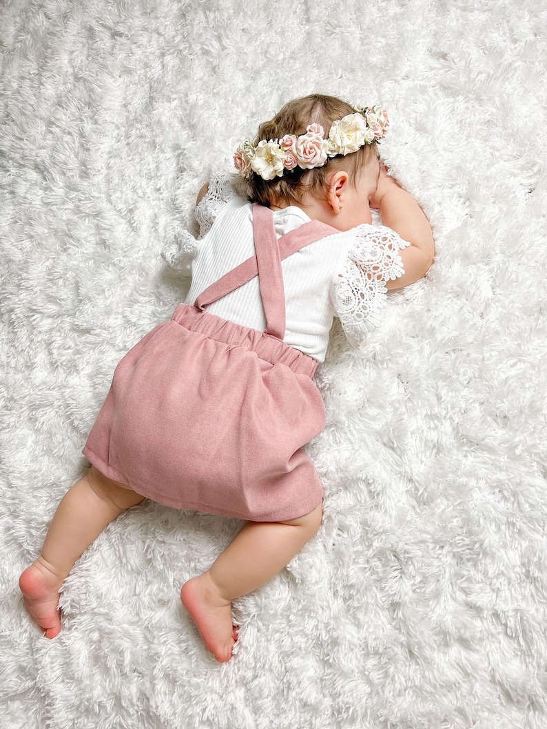 May include: A baby girl wearing a pink skirt and a white shirt with straps. She is wearing a flower crown and is lying on a white fluffy rug.