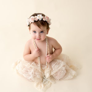 May include: A baby girl wearing a white lace dress and a flower crown, holding a pearl necklace.