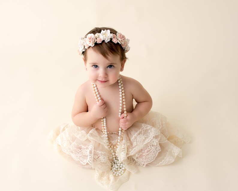 May include: A baby girl wearing a white lace dress and a flower crown, holding a pearl necklace.