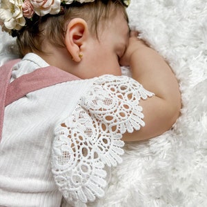 May include: A baby girl wearing a white lace dress with a pink bodice and a flower crown. The baby is sleeping on a white fluffy blanket.