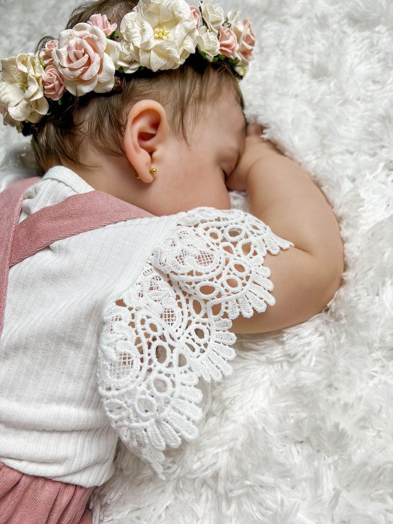 May include: A baby girl wearing a white lace dress with a pink bodice and a flower crown. The baby is sleeping on a white fluffy blanket.