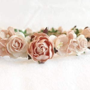 May include: A delicate floral crown with blush pink paper flowers and green leaves. The crown is perfect for a wedding, bridal shower, or other special occasion.