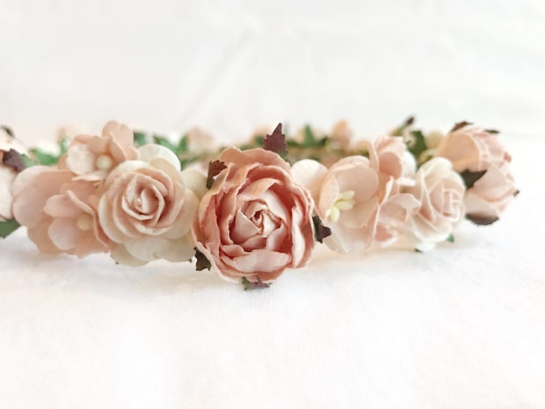 May include: A delicate floral crown with blush pink paper flowers and green leaves. The crown is perfect for a wedding, bridal shower, or other special occasion.