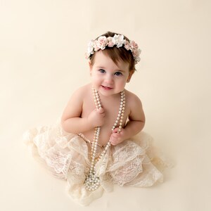 May include: A baby girl wearing a white lace skirt and a flower crown, holding a pearl necklace.