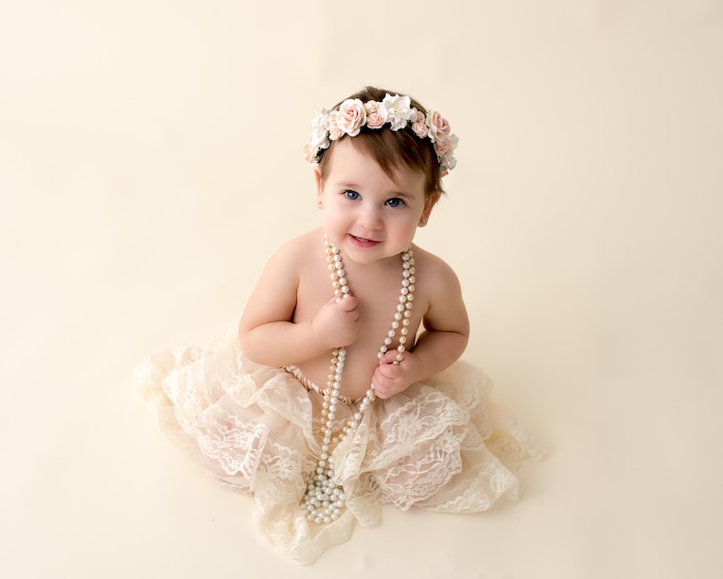 May include: A baby girl wearing a white lace skirt and a flower crown, holding a pearl necklace.