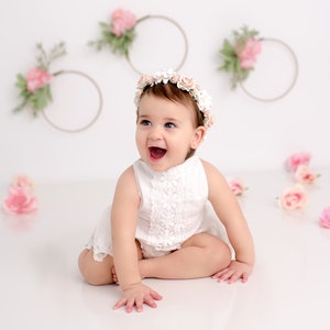 May include: A baby girl wearing a white lace dress and a flower crown smiles at the camera. The background is white with pink flowers and wreaths.