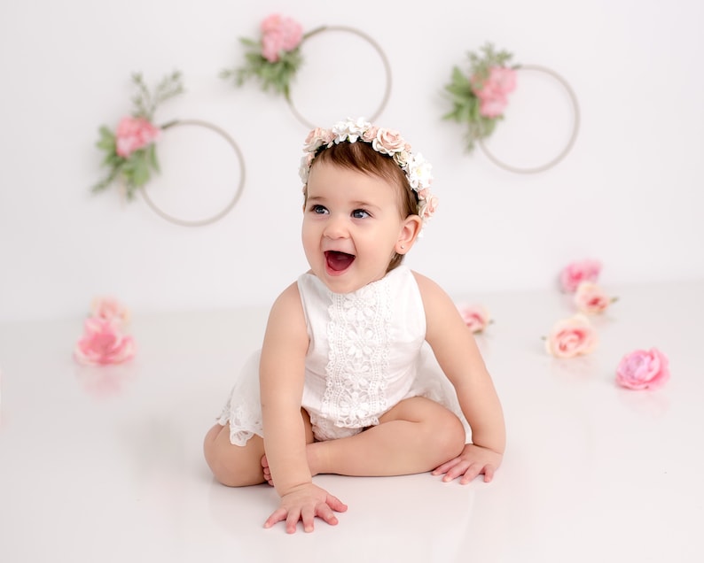 May include: A baby girl wearing a white lace dress and a flower crown smiles at the camera. The background is white with pink flowers and wreaths.