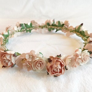 May include: A delicate floral crown with blush pink and white paper flowers and green leaves. The crown is made of a woven wire base.