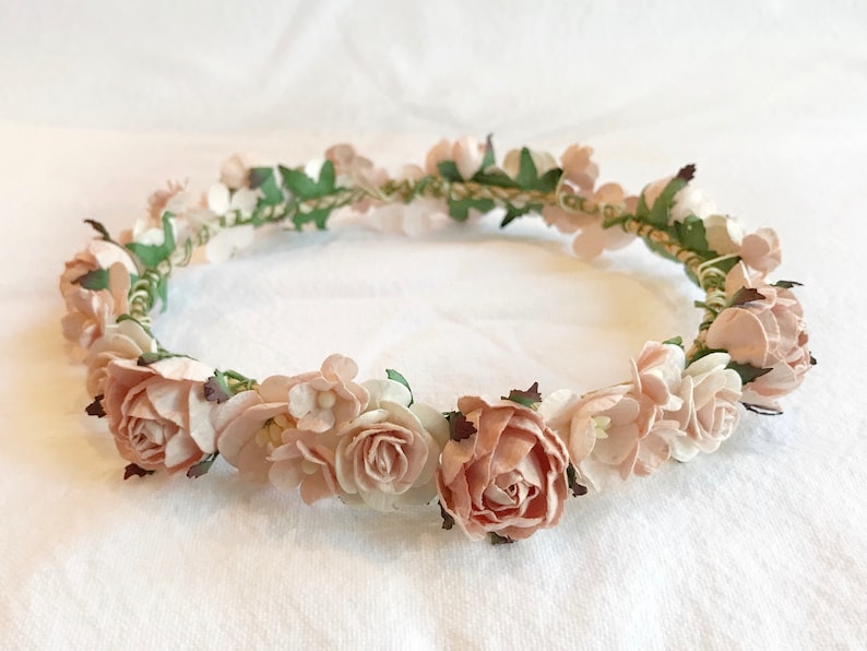 May include: A delicate floral crown with blush pink and white paper flowers and green leaves. The crown is made of a woven wire base.