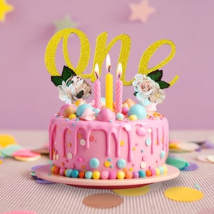 May include: A pink cake with colorful sprinkles and a gold glitter "one" topper. Three lit candles are on top of the cake.