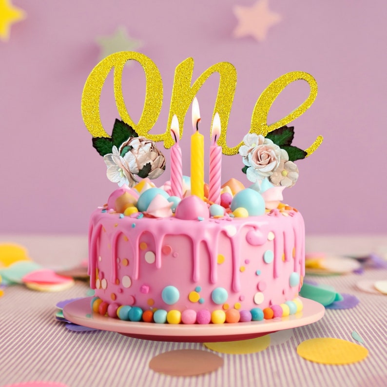 May include: A pink cake with colorful sprinkles and a gold glitter "one" topper. Three lit candles are on top of the cake.