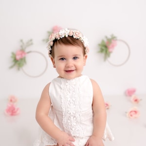 May include: A baby girl wearing a white dress with lace trim and a flower crown smiles at the camera. The background is white with pink flowers.