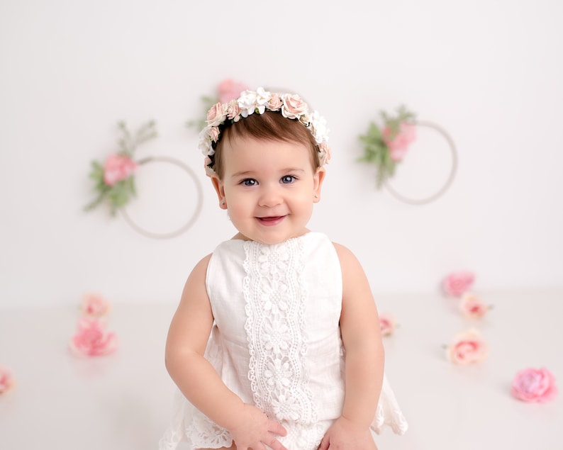 May include: A baby girl wearing a white dress with lace trim and a flower crown smiles at the camera. The background is white with pink flowers.