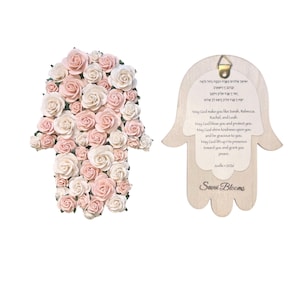 May include: Two Hamsa hand-shaped wall hangings. One is covered in pink and white paper roses. The other is made of light wood with a printed blessing in English and Hebrew. The wooden Hamsa has the text "Savvi Blooms".