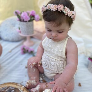 May include: A baby girl wearing a white lace dress and a pink flower crown is sitting on a white blanket and eating a cake with frosting on her face and hands.
