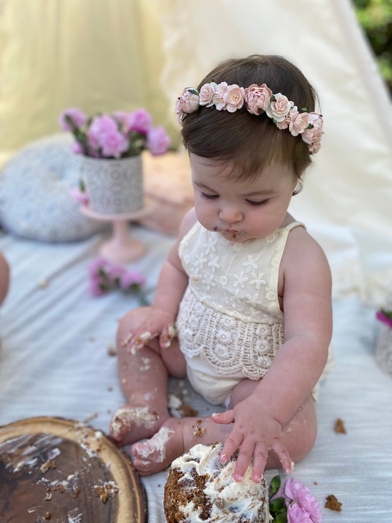 May include: A baby girl wearing a white lace dress and a pink flower crown is sitting on a white blanket and eating a cake with frosting on her face and hands.