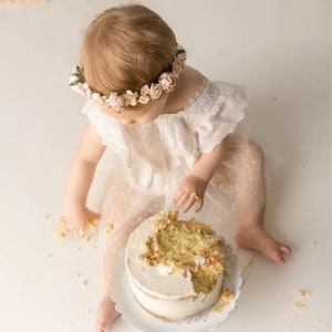 May include: A baby girl wearing a white dress with a floral crown is sitting in front of a white cake with a bite taken out of it.