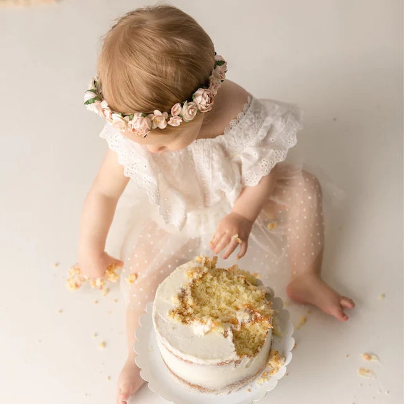 May include: A baby girl wearing a white dress with a floral crown is sitting in front of a white cake with a bite taken out of it.