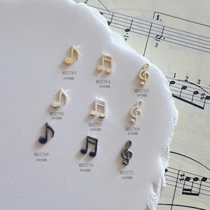 May include: A collection of musical note charms in gold, silver, and black. The charms include eighth notes, quarter notes, and treble clefs. Sizes range from 4mm x 9mm to 6mm x 8mm. The background includes sheet music.